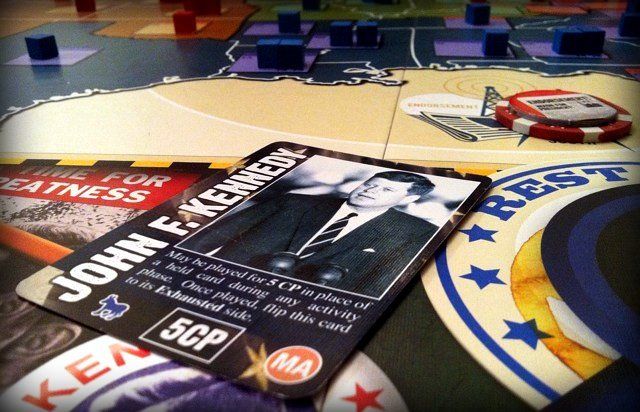 A close-up of GMT Games' "1960: The Making of the President (Second Printing)" board game displays a card featuring John F. Kennedy, various game pieces, and sections of the 1960 election-themed board in the background.