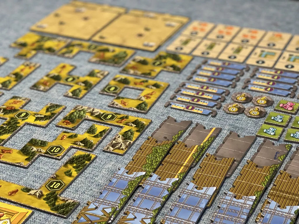 A close-up of colorful components from Lookout Games' "Bärenpark: The Bad News Bears Expansion," featuring bridge sections, grizzly bear tiles, tokens, and terrain tiles neatly arranged on a blue-gray surface.