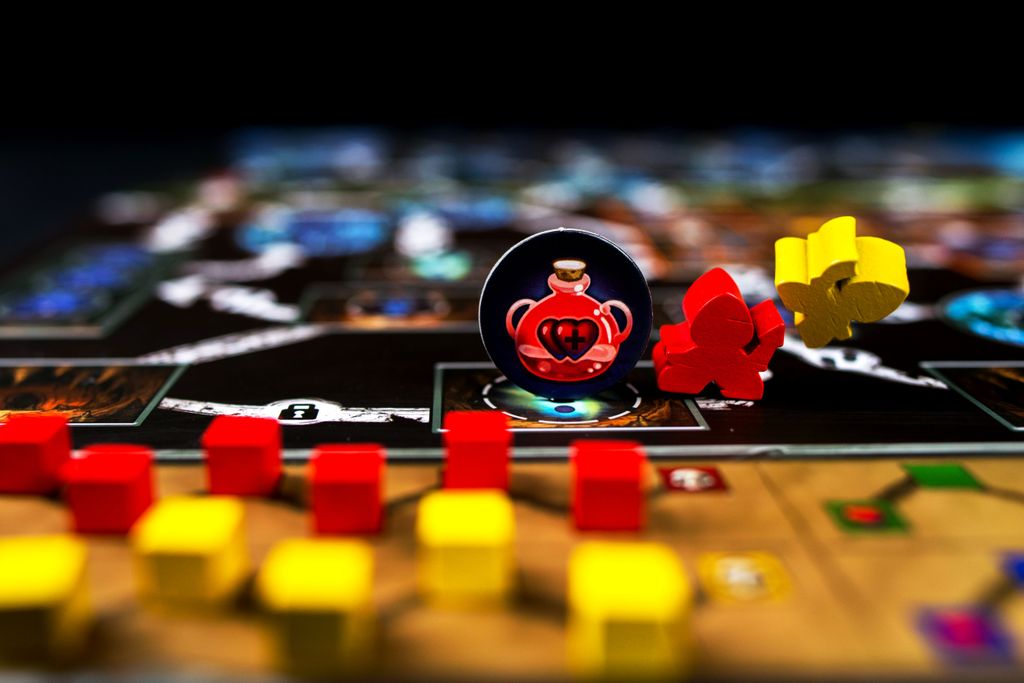 A close-up of Clank!: A Deck-Building Adventure by Renegade Game Studios, showing red and yellow wooden pieces, a red potion token, and a red meeple on a vibrant board with paths to the dragon’s lair. Background is blurred.