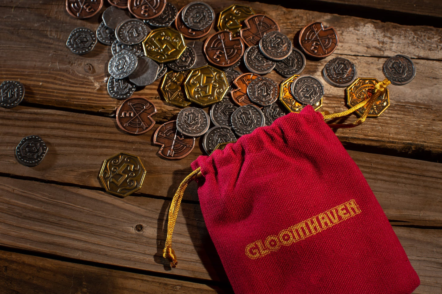 A red Gloomhaven bag by Cephalofair Games spills premium gold, bronze, and silver Gloomhaven: Metal Coin Upgrade tokens across a rustic wooden table, adding style and quality to your game.