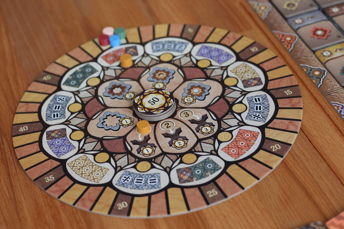 The Intarsia board game by Pegasus Spiele features a colorful, circular design with intricate intarsia patterns, numbered spaces, tokens, and pieces set on a wooden table. A similarly styled rectangular section is also included nearby.