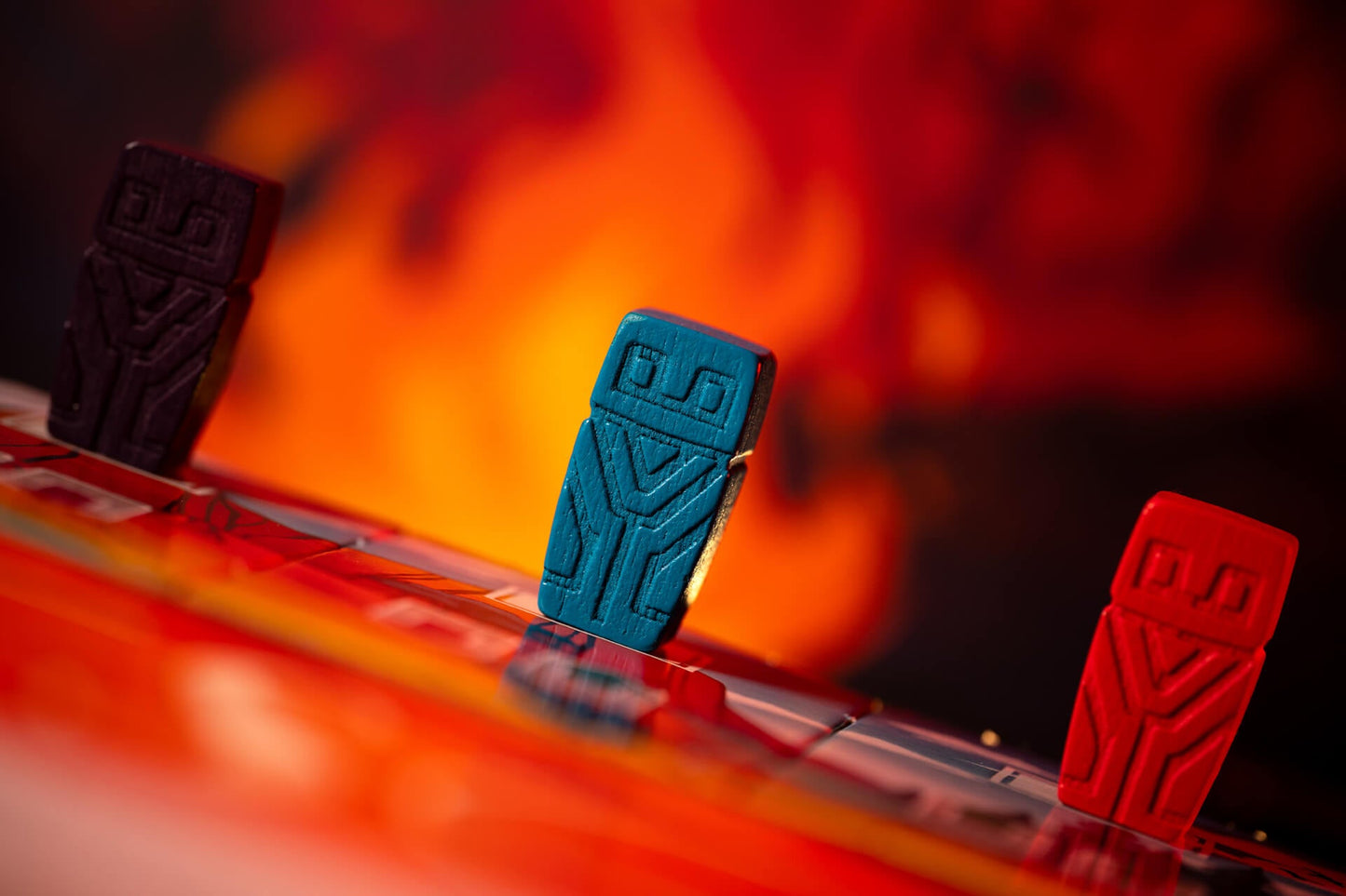 Three colorful game pieces—purple, blue, and red—from Pegasus Spiele's Mesos stand on a reflective board with a fiery abstract backdrop, evoking the intense strategy and tribal competition central to this unique game.