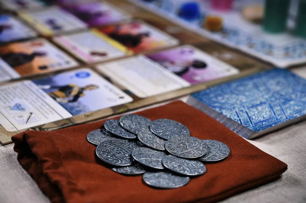 A set of ornate Pax Pamir: Second Edition Metal Coins & Cloth Bag by Wehrlegig Games is displayed next to a deck of blue patterned cards and illustrated game cards, evoking a classic Pax Pamir tabletop setup.
