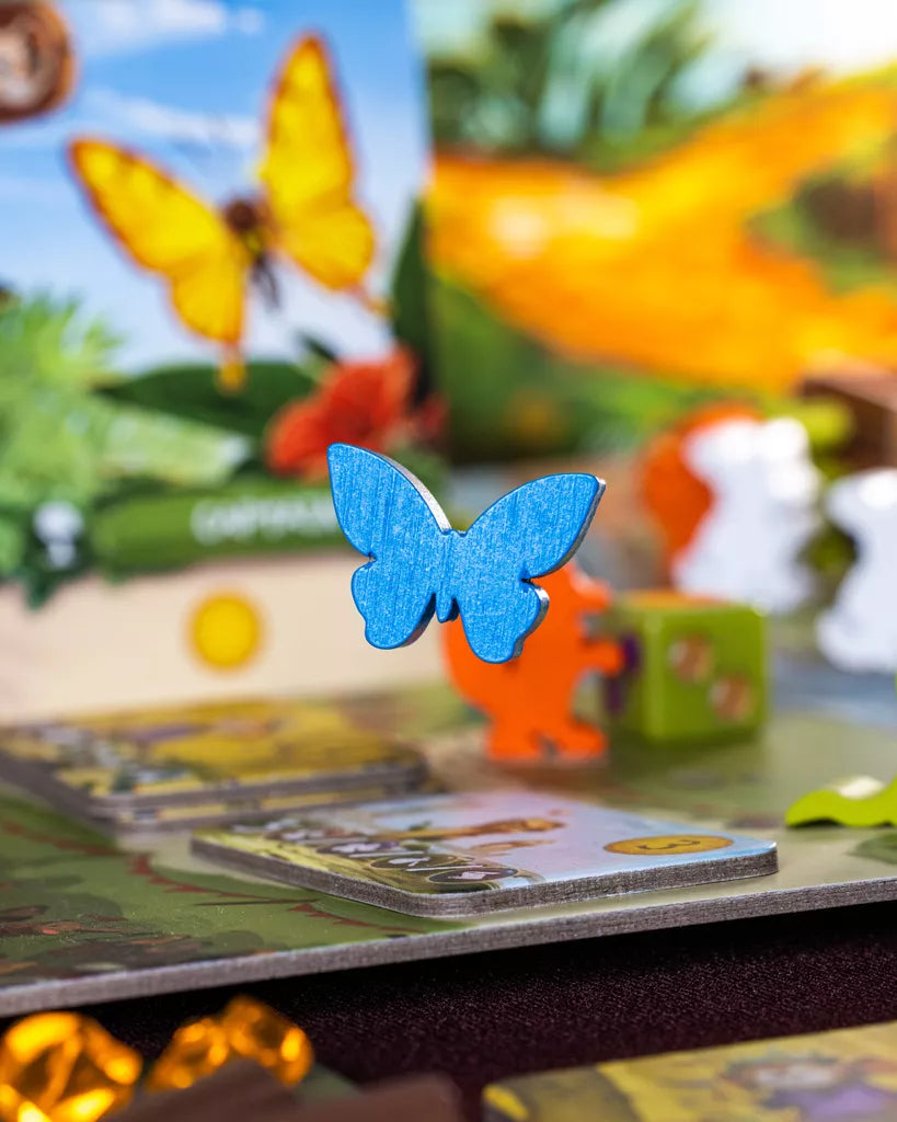 A close-up of a blue butterfly-shaped piece on the vibrant My Lil' Everdell board game by Starling Games (II), with cards, tokens, and a yellow butterfly illustration visible in the blurred background.