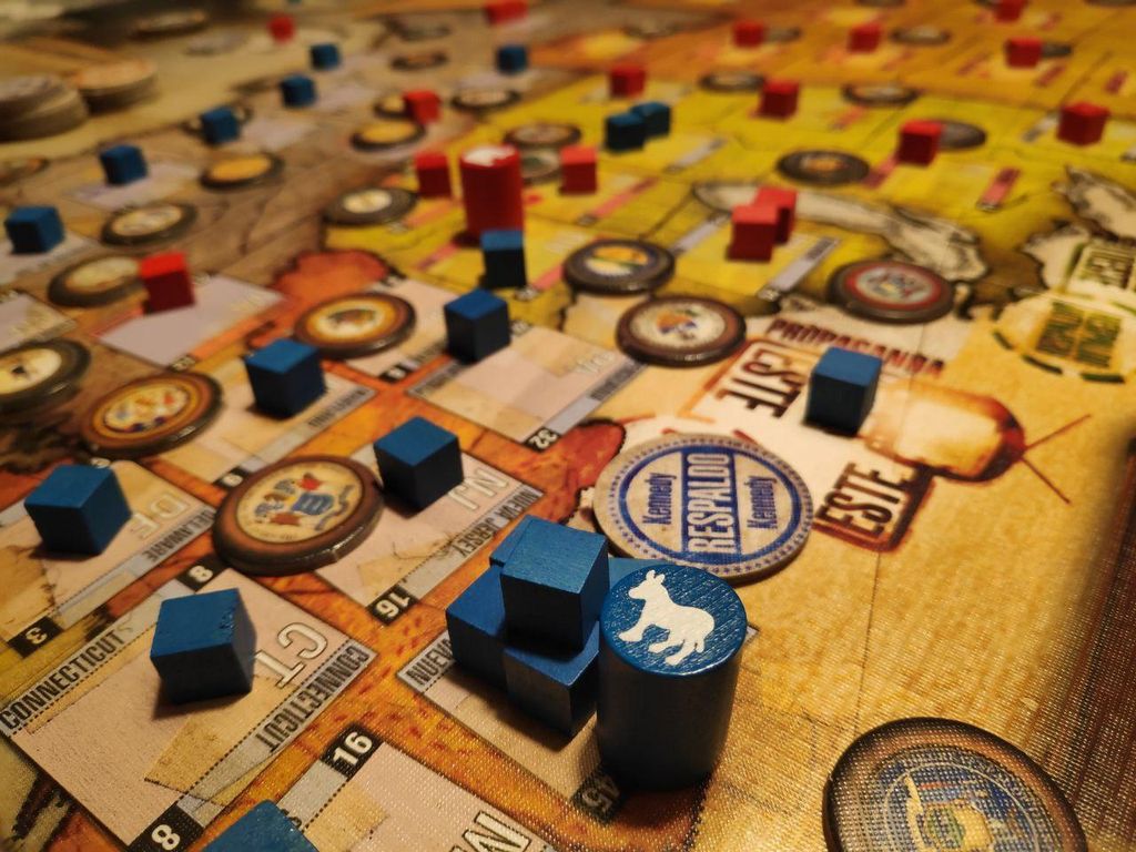 A close-up of 1960: The Making of the President (Second Printing) by GMT Games in play, showing blue cubes and discs with white animal symbols and red cubes on a board depicting regions from the historic Kennedy vs Nixon election.