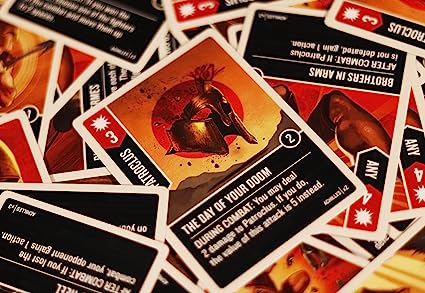 A stack of illustrated playing cards from Unmatched: Battle of Legends, Volume Two (Iello), with the central card titled The Day of Your Doom showing a helmeted warrior in bold red and orange, surrounded by more vibrant game cards.
