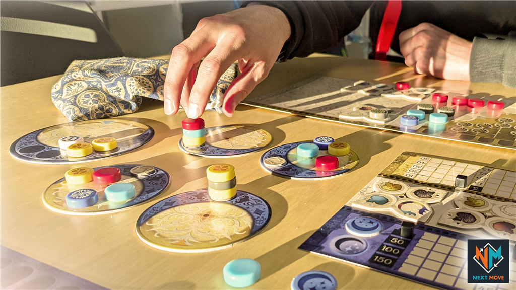 Two people play Azul Duel NL/FR by Next Move Games, placing colorful discs on circular tiles. One hand sets a red disc as tokens and a score sheet are on the wooden table, illuminated by sunlight.
