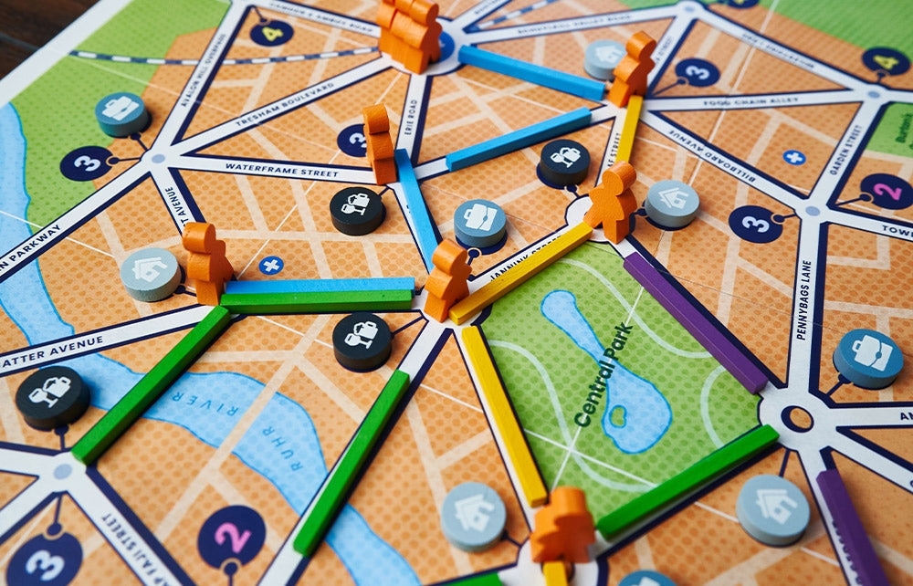 A close-up of Bus: Complete Edition by Capstone Games features orange wooden pieces, multicolored route sticks, and icon tokens, all set on a map showing “Central Park” and street names.