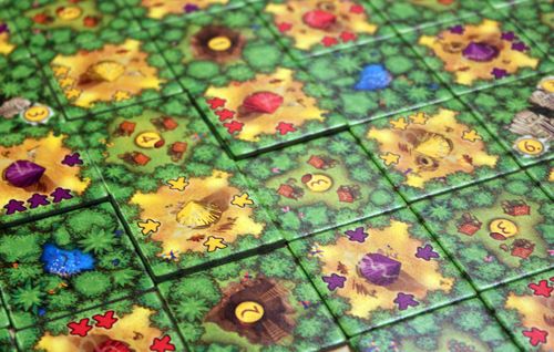 A close-up of Cacao NL tiles from White Goblin Games shows green forests, numbered yellow clearings, colored huts, and icons for animals or resources scattered across the vibrant tile-placement board game.