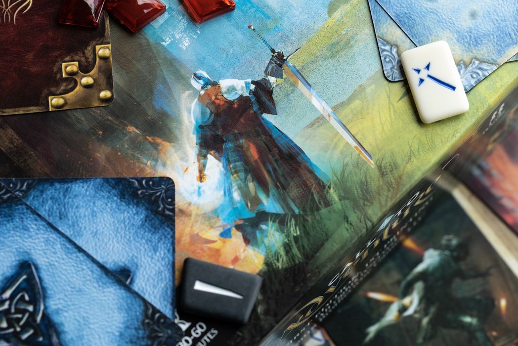 A close-up of Brotherwise Games’ Call to Adventure shows illustrated cards, a knight with a sword, red tokens, a white tile with a blue symbol, and a black token on the colorful game board.