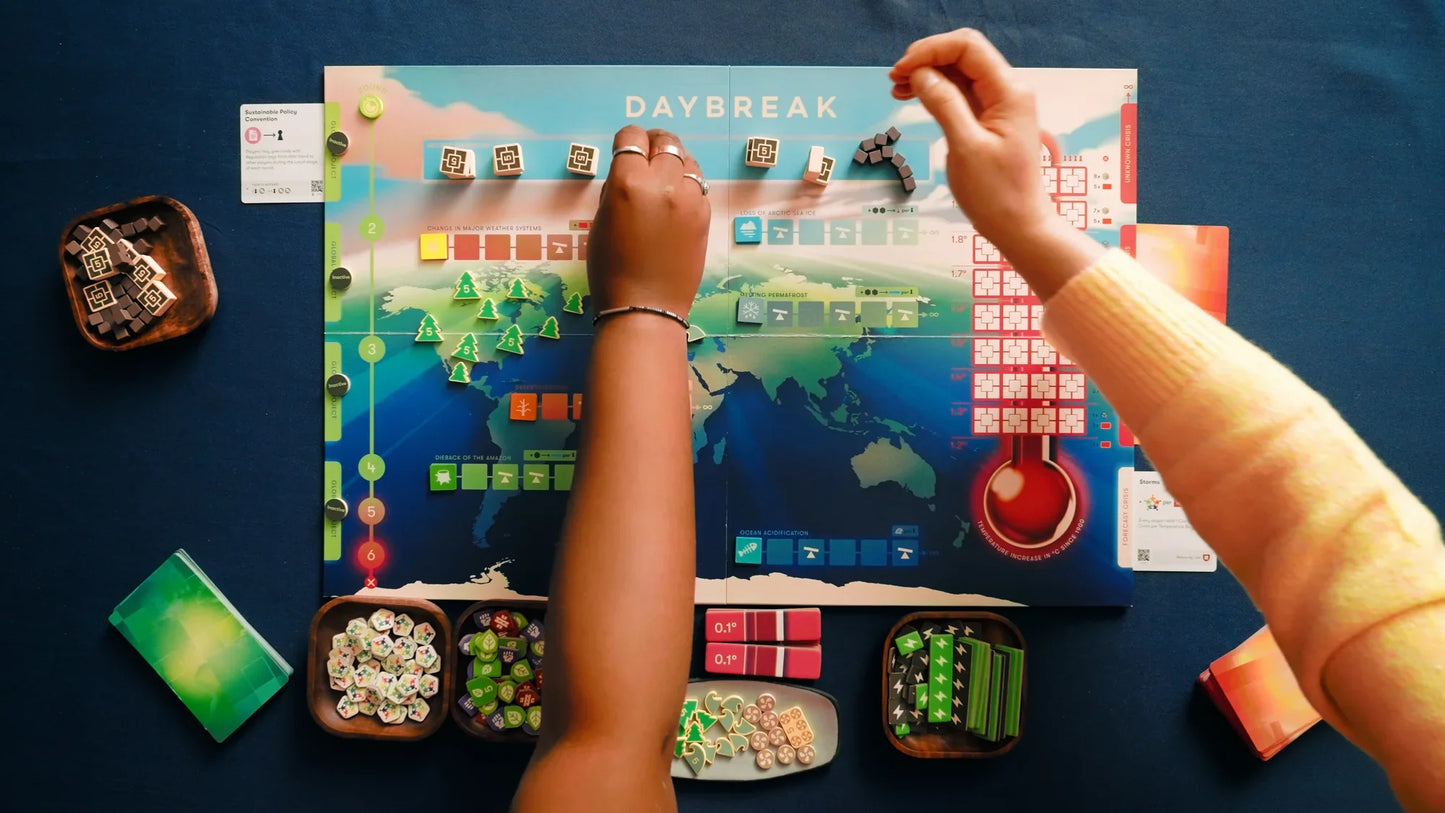 Two people play CMYK’s cooperative game Daybreak, placing pieces on its colorful map. Game tokens, cards, and wooden bowls filled with pieces are neatly set out on a blue surface.