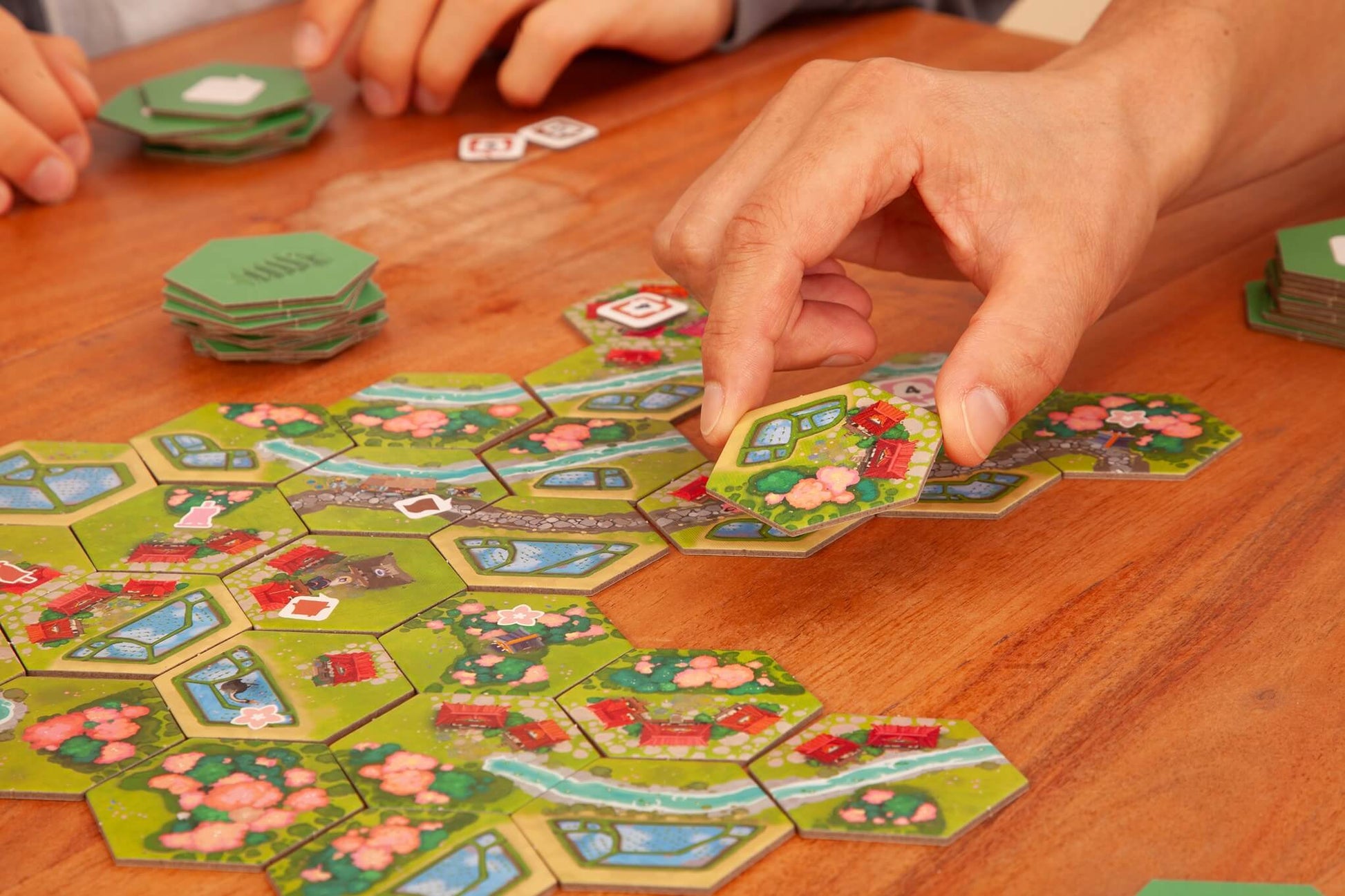 A close-up of hands playing Pegasus Spiele’s Dorfromantik: Sakura, placing hexagonal tiles with landscapes, trees, and cherry blossoms on a wooden table, while other tiles from the game are stacked nearby.