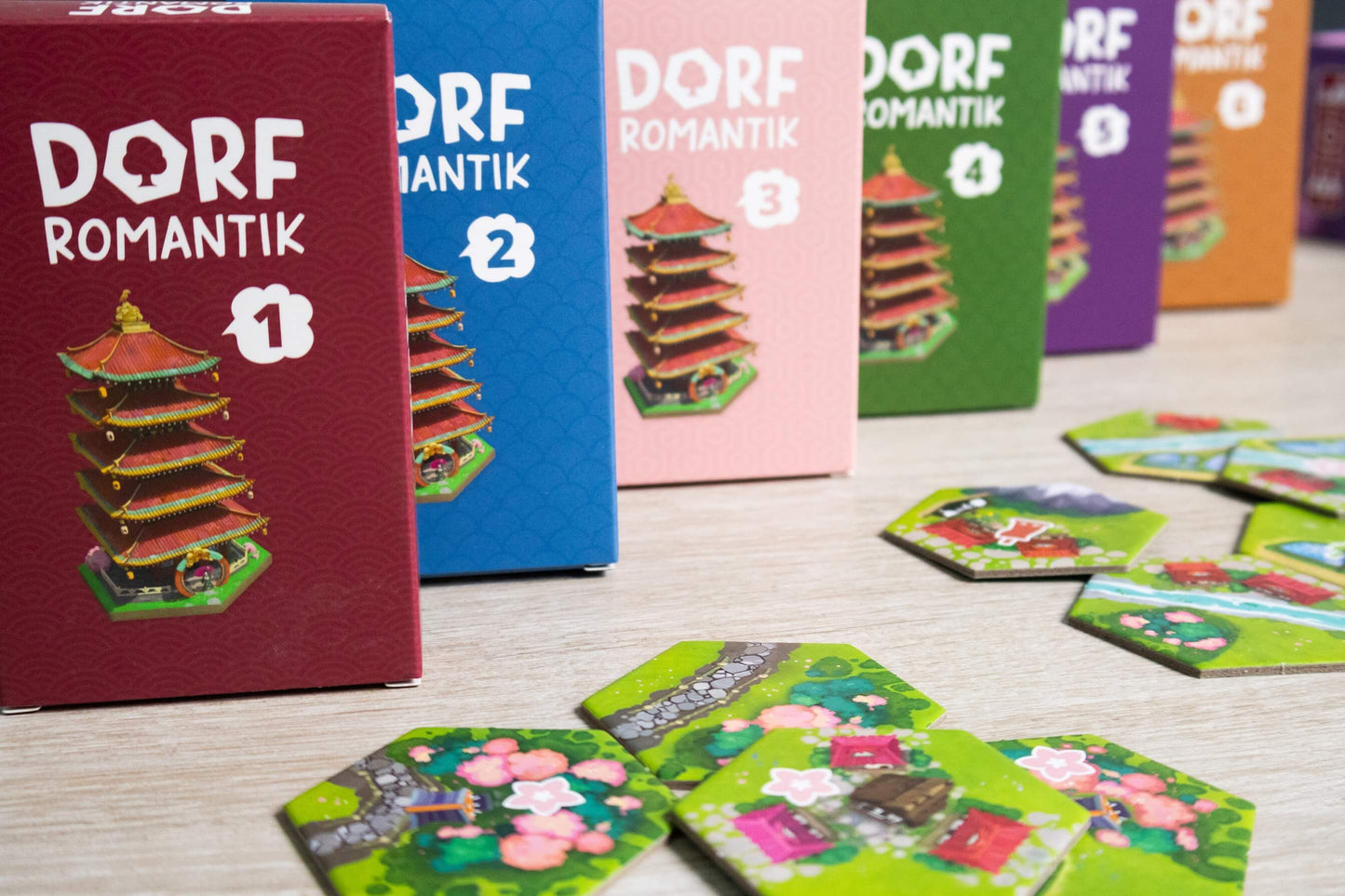 Colorful boxes labeled “Dorfromantik” (1–5) from Pegasus Spiele stand in a row. Hexagonal tiles, including cherry blossoms from Dorfromantik: Sakura, are scattered on a light wood surface in front.