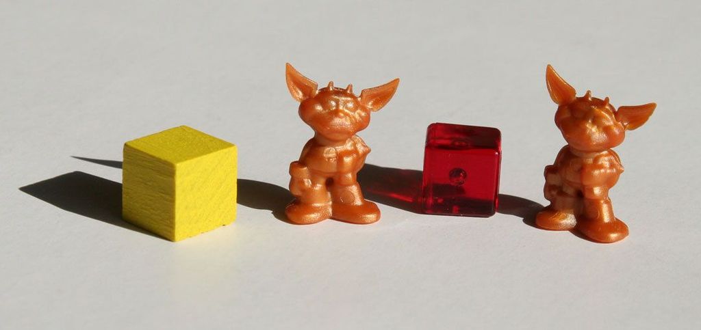 Two small orange goblin figurines from Dungeon Lords by Czech Games Edition (CGE) stand side by side on a white surface, flanked by a yellow cube to the left and a translucent red cube between them.