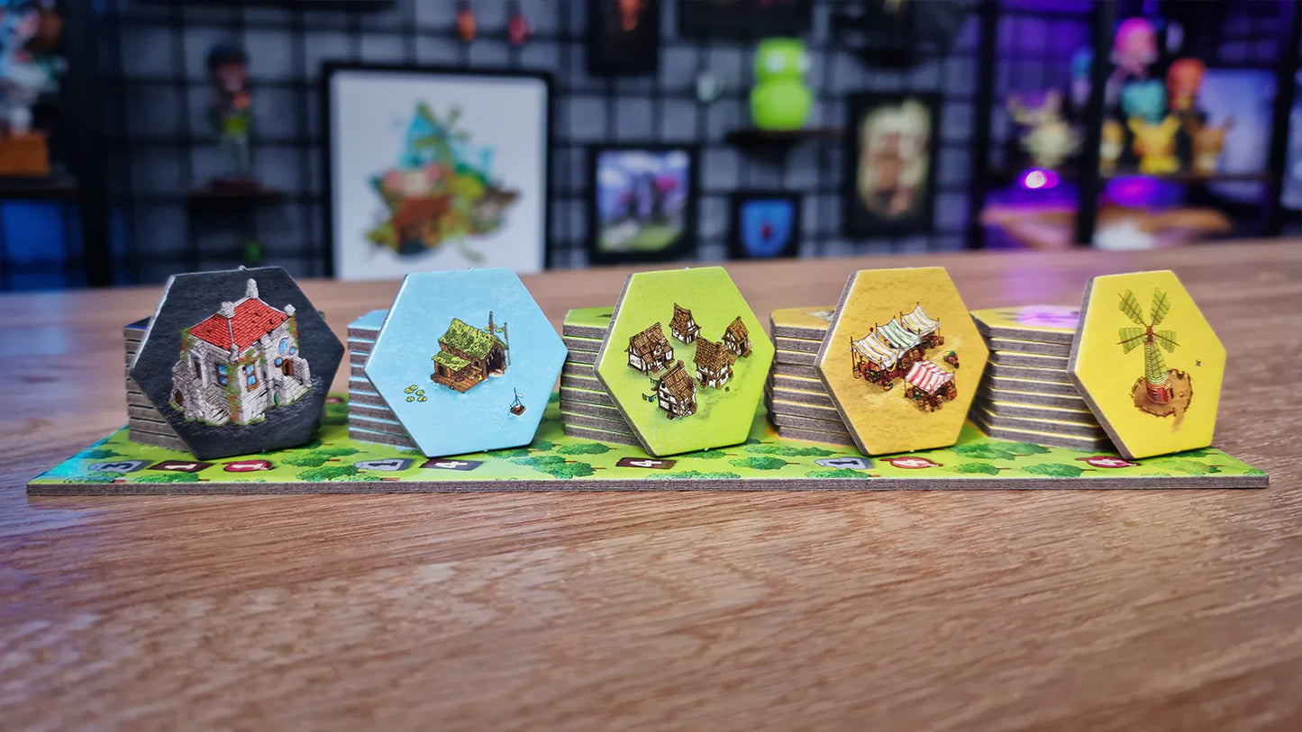 Four stacks of hexagonal tiles from Emberleaf by The City of Games, featuring buildings, houses, and a windmill, sit on a wooden table with shelves and artwork blurred in the colorful background.