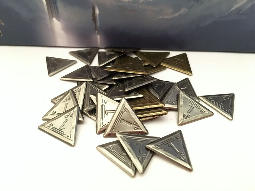 Metallic triangular tokens from Empires of the Void II by Red Raven Games, featuring engraved markings such as "1," appear in silver, gold, and dark gray, scattered on a white surface with a blurred background.