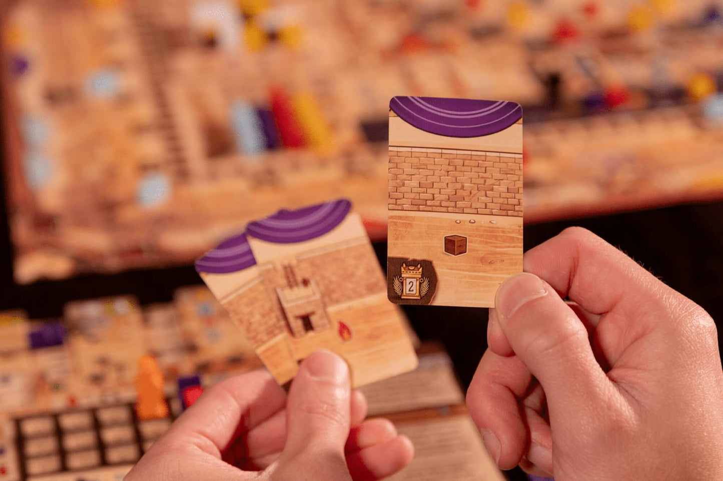 Two hands hold up Ezra and Nehemiah cards by Garphill Games, featuring brick wall and purple curtain designs with a box and lock, set against a blurred board game scene with pieces in the background.