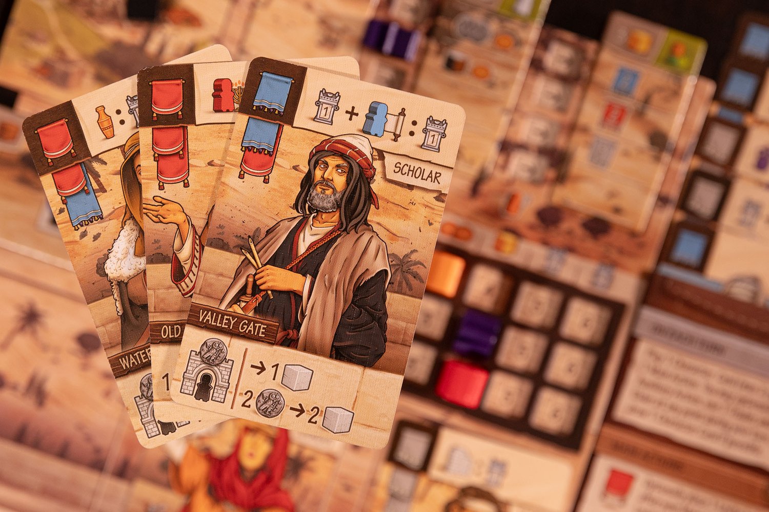 Three illustrated playing cards from "Ezra and Nehemiah" by Garphill Games—featuring historical figures and symbols—are held above a tabletop filled with cards, cubes, and other strategy board game components.