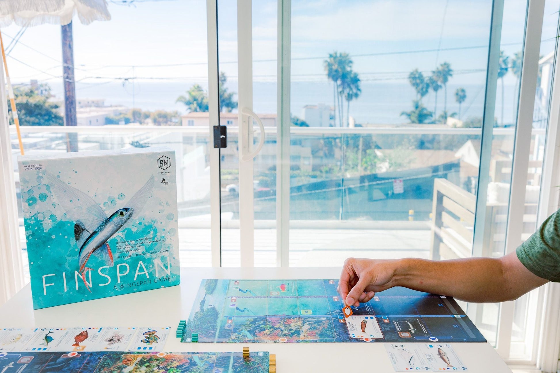 A person plays Finspan by Stonemaier Games at a white table near sunny windows with ocean and palm trees in view. The Finspan game box is on the table, ideal for marine researchers or fans of aquatic life.