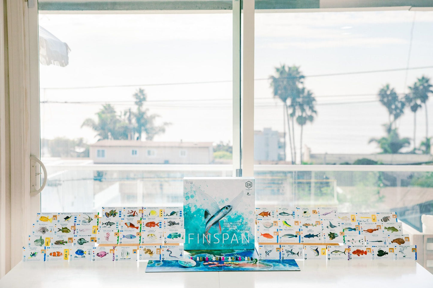 Finspan by Stonemaier Games is displayed on a table by a window with views of palm trees and the ocean. Behind the main box, colorful cards feature aquatic creatures from various ocean zones.