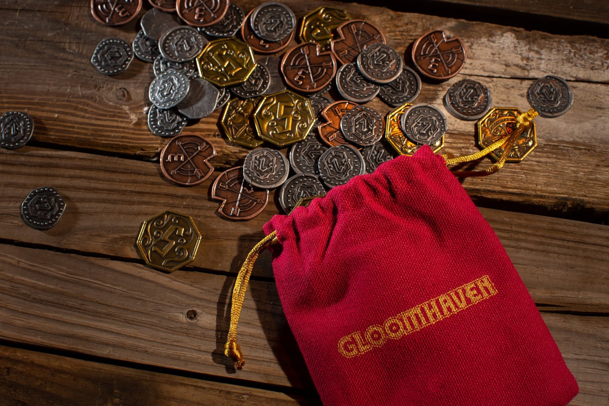 A red Gloomhaven bag by Cephalofair Games spills premium gold, bronze, and silver Gloomhaven: Metal Coin Upgrade tokens across a rustic wooden table, adding style and quality to your game.