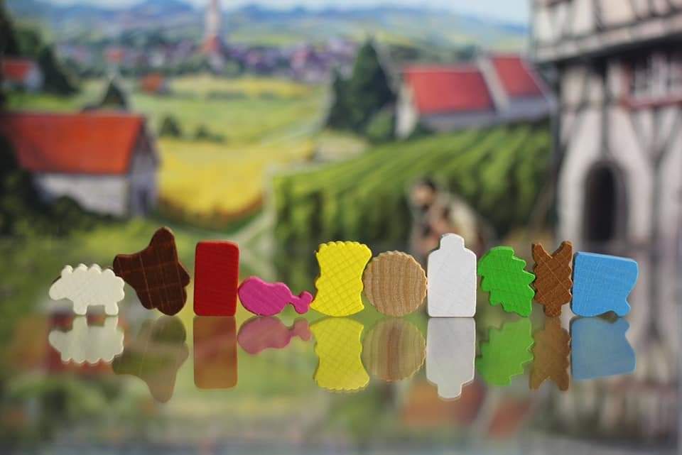 Vibrant wooden game tokens shaped like animals and objects from Lookout Games’ Hallertau are displayed on a reflective surface, capturing its agricultural and sheep-breeding themes with a blurred countryside village backdrop.