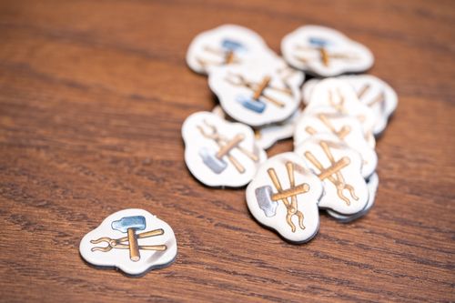 A pile of small, cloud-shaped Hallertau tokens from Lookout Games, showing crossed hammer and tongs, rests on a wooden surface. Ideal for enhancing worker placement in this agriculture and sheep breeding game. One token is separated.