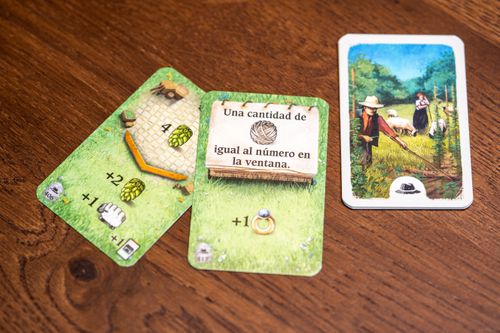 Three illustrated Hallertau game cards by Lookout Games lie on a wooden table: two display resources and icons, including text "Una cantidad de igual al número en la ventana," while the third depicts people in agriculture and sheep breeding.