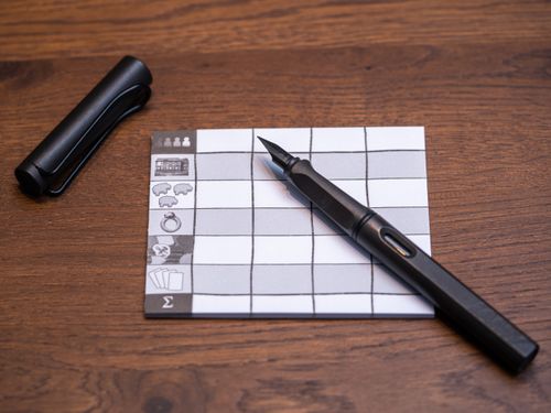 A black fountain pen sits on a small grid notepad featuring icons, reminiscent of planning in Lookout Games' Hallertau board game, all arranged atop a wooden surface with the pen cap placed above the pad.