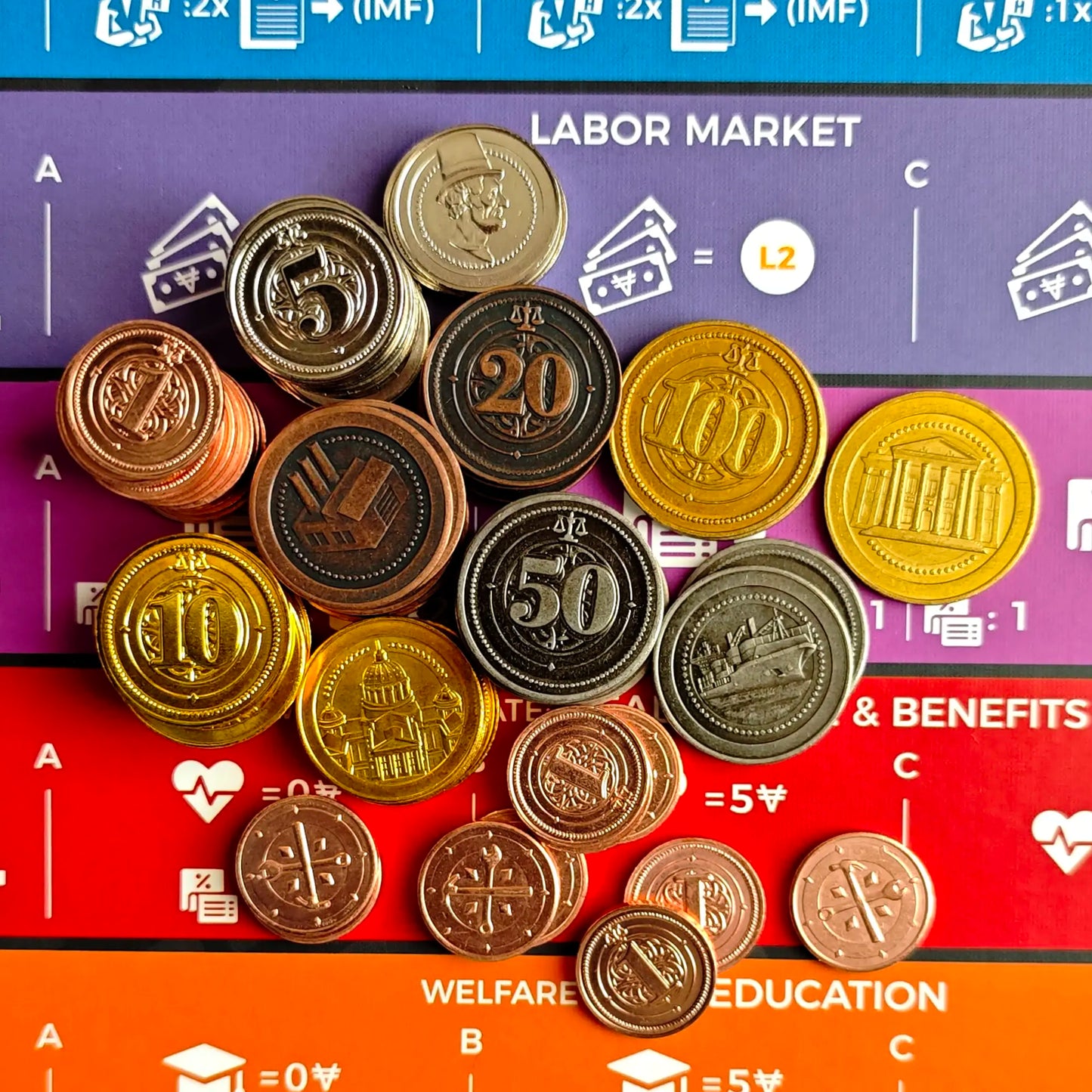 Hegemonic Project Games’ Hegemony: Deluxe Metal Coins in assorted sizes and values are displayed across a board labeled with categories such as “LABOR MARKET,” “TAXES & BENEFITS,” and “WELFARE & EDUCATION.”.