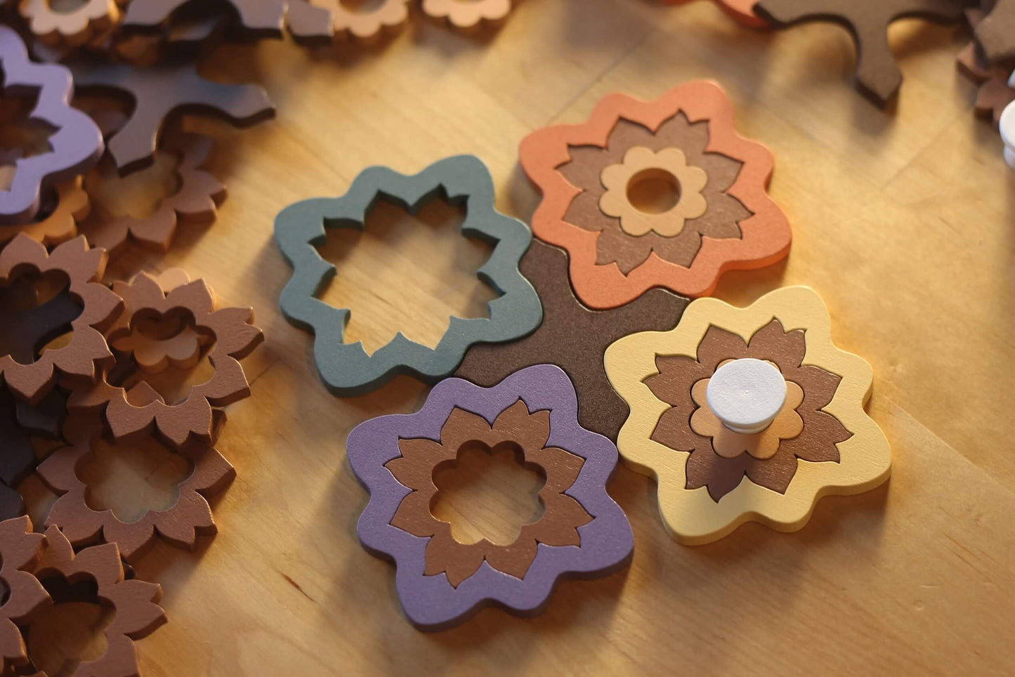 Four vibrant, flower-shaped wooden pieces from the Intarsia board game by Pegasus Spiele interlock on an artisan surface, with more pieces scattered nearby. A yellow piece features a white circular peg at its center, all under warm lighting that highlight