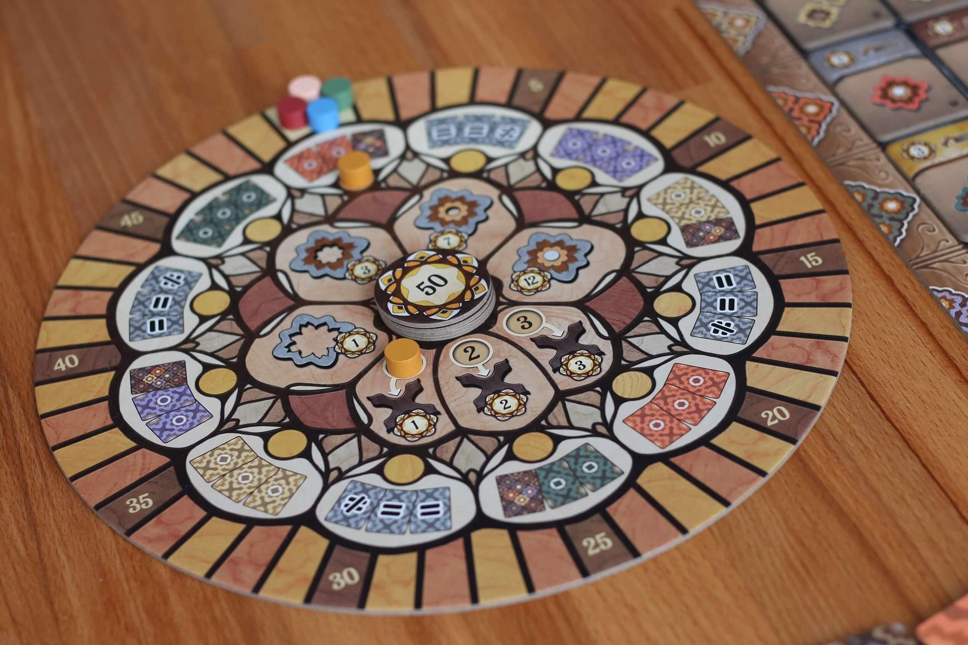 The Intarsia board game by Pegasus Spiele features a colorful, circular design with intricate intarsia patterns, numbered spaces, tokens, and pieces set on a wooden table. A similarly styled rectangular section is also included nearby.
