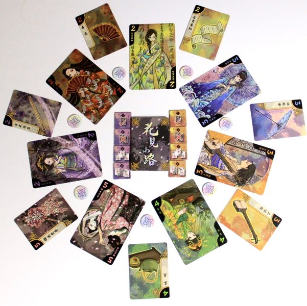 A circular flat lay displays Hanamikoji playing cards by EmperorS4, featuring Japanese art and floral themes. Small flower tokens are scattered around a central card on a white background, evoking the aesthetic of the Hanamikoji game.