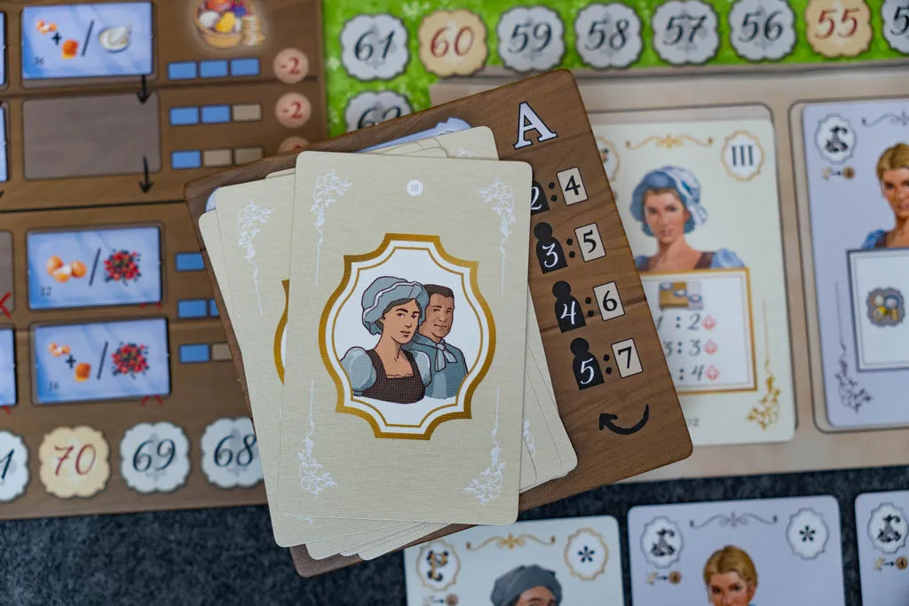 A close-up of La Pâtisserie Rococo by Eagle-Gryphon Games shows illustrated cards with people in period attire, a wooden scoring board with numbers and a smiley face, plus various components, reflecting the elegance of the Versailles Garden Festival.