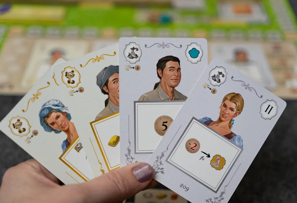 A hand displays four illustrated playing cards from Eagle-Gryphon Games’ La Pâtisserie Rococo, each featuring artwork inspired by the game. In the background is a blurred game board, evoking a Versailles garden festival.