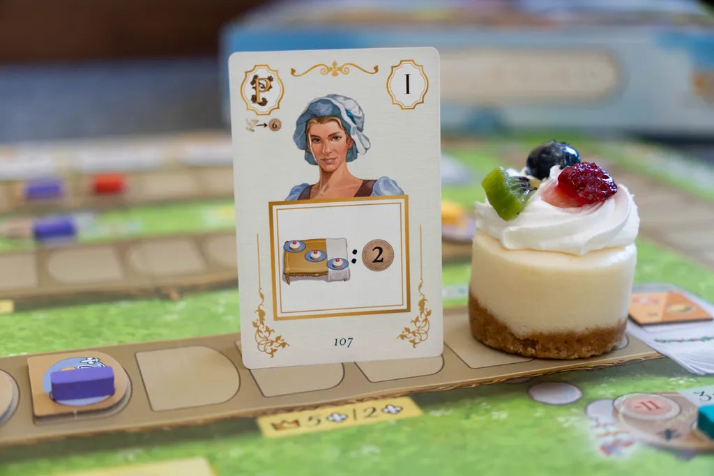 A card from La Pâtisserie Rococo by Eagle-Gryphon Games, showing a woman in a bonnet and desserts, stands on the board next to a fruit-topped cheesecake, capturing the elegance of pastry chefs at Versailles.