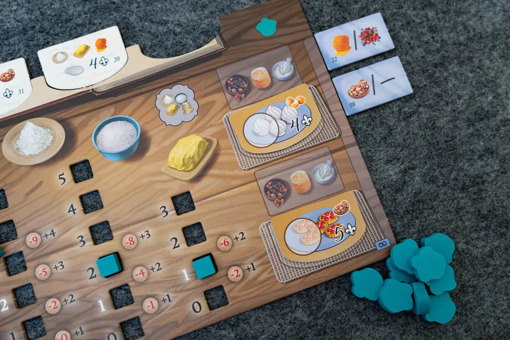 A close-up of La Pâtisserie Rococo by Eagle-Gryphon Games shows detailed food illustrations—flour, butter, eggs, and pancakes with toppings. Blue tokens are placed on the board, which sits on a gray felt surface, evoking a pastry chef's creativity.
