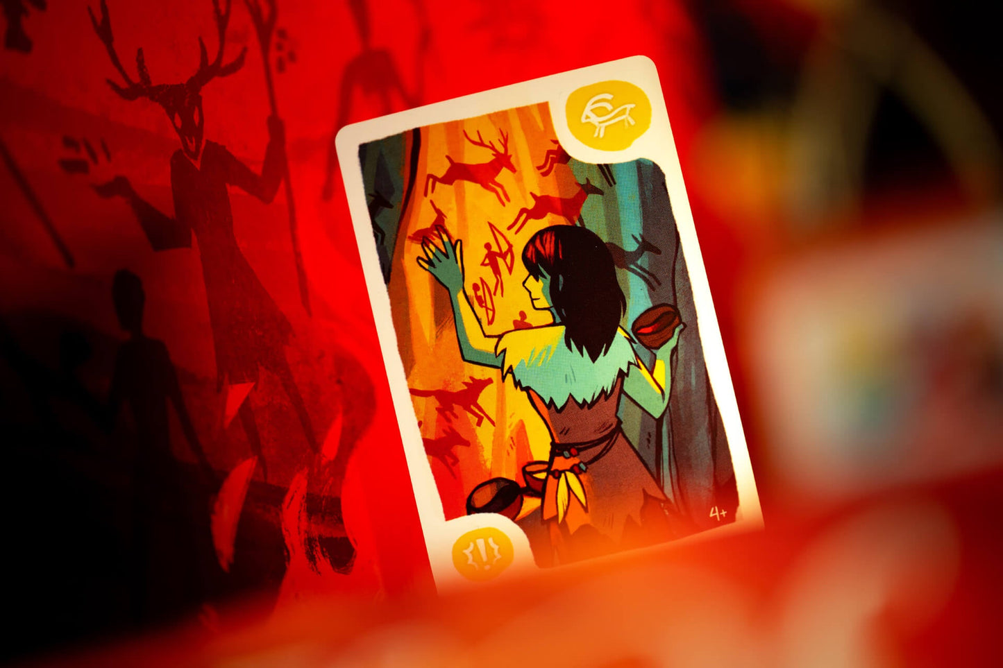 The Mesos playing card from Pegasus Spiele depicts a dark-haired figure in fur clothing painting ancient symbols on a cave wall, set against a red, shadowy background with figures inspired by prehistoric art.
