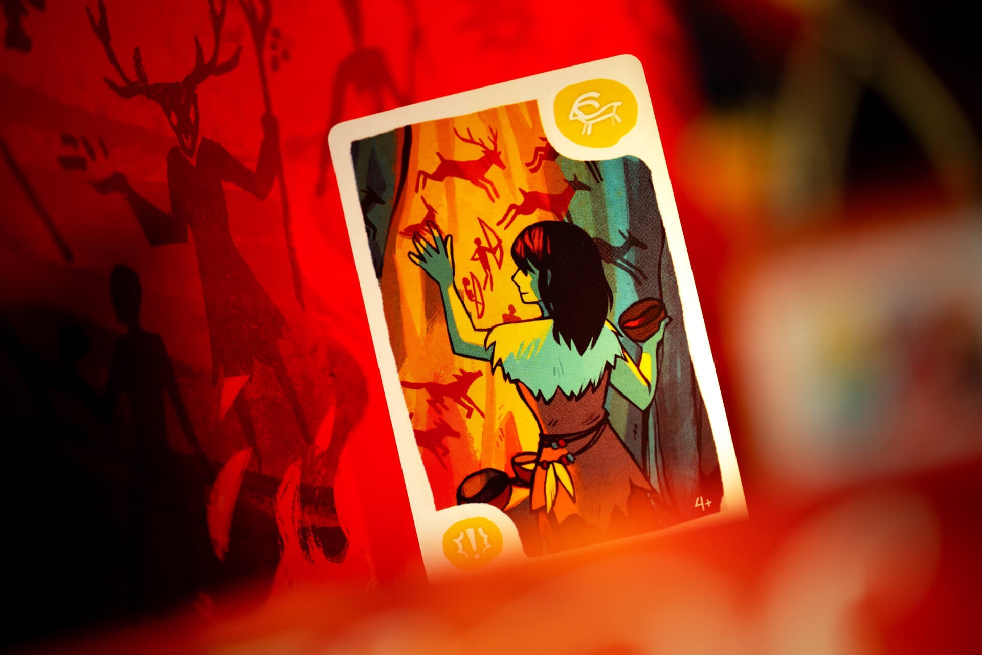 The Mesos playing card from Pegasus Spiele depicts a dark-haired figure in fur clothing painting ancient symbols on a cave wall, set against a red, shadowy background with figures inspired by prehistoric art.