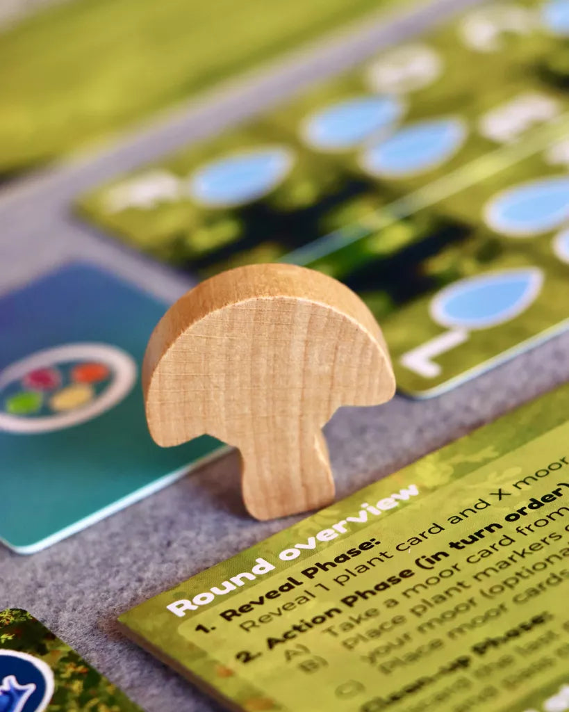 A wooden mushroom-shaped piece from Moorland by Pegasus Spiele stands on a table next to colorful cards and a Round Overview rule sheet, highlighting the game's ecosystem management theme. A softly blurred background draws focus to the components.