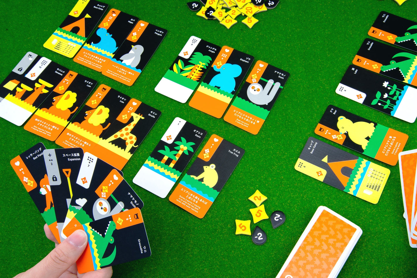 On a green table, Oink Games' "Moving Wild" features animal-illustrated cards and yellow diamond tokens. A hand holds five cards, while more cards and tokens are spread across the play area in this colorful drafting game.