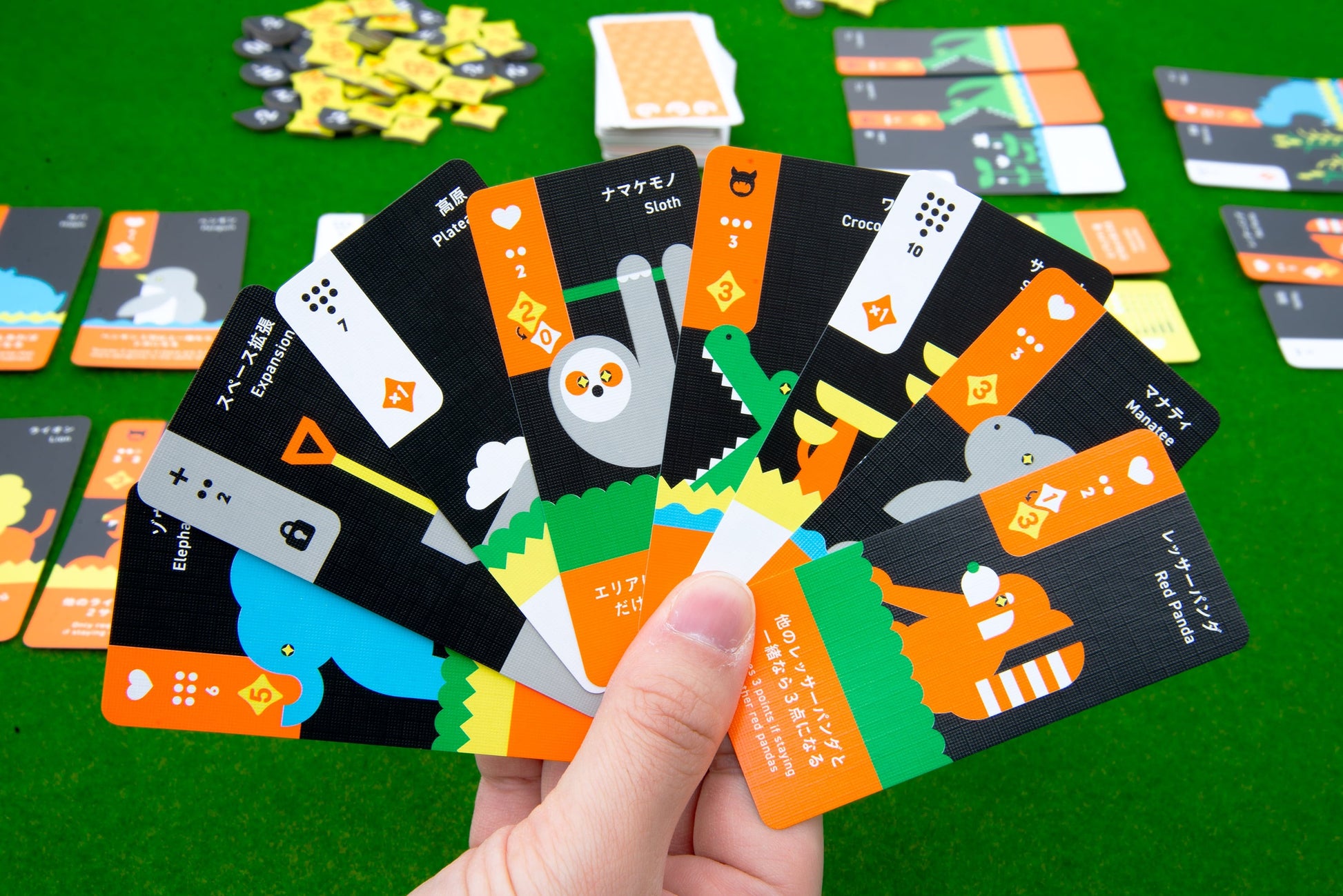 A hand holds seven colorful Moving Wild cards from Oink Games, each with cartoon animals and numbers, for a drafting game about animal migration; more cards and pieces are spread out on a green surface in the background.