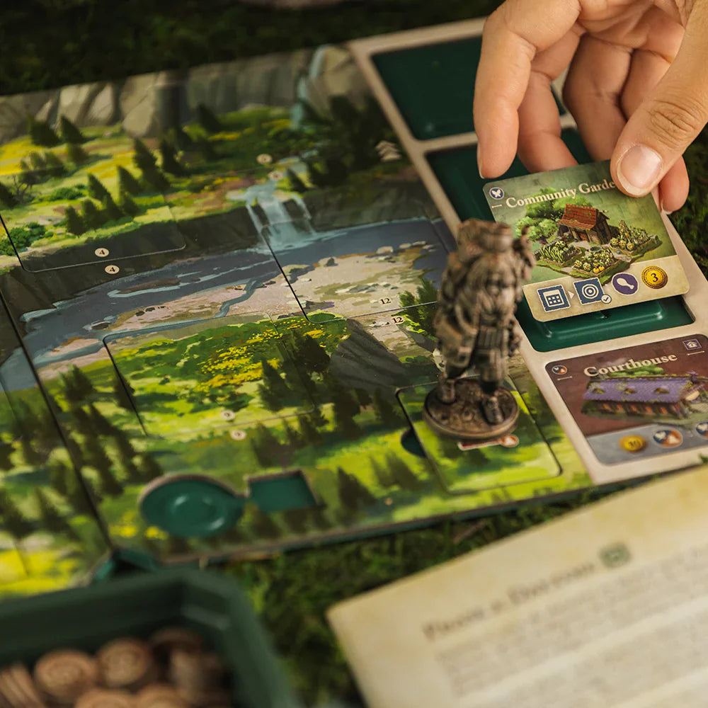 A hand places a Community Garden card on the colorful Mythwind (Damaged) board by Open Owl Studios. The landscape, figurine, and nearby scattered game pieces suggest this cooperative board game is currently being played.