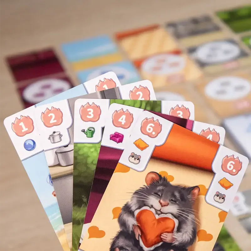 A close-up of a hand holding four illustrated Paws Up cards from Pegasus Spiele, showing icons, numbers, and a cute gray hamster with a heart-shaped object. Colorful cards and game pieces appear blurred in the background.
