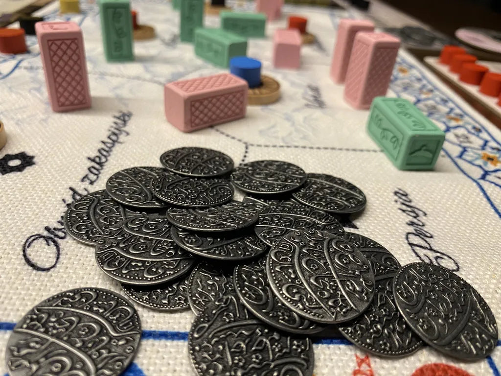 Close-up of Wehrlegig Games' Pax Pamir: Second Edition Metal Coins & Cloth Bag, featuring ornate coins on a patterned board with colorful wooden pieces and a partially visible embroidered cloth bag in the background.
