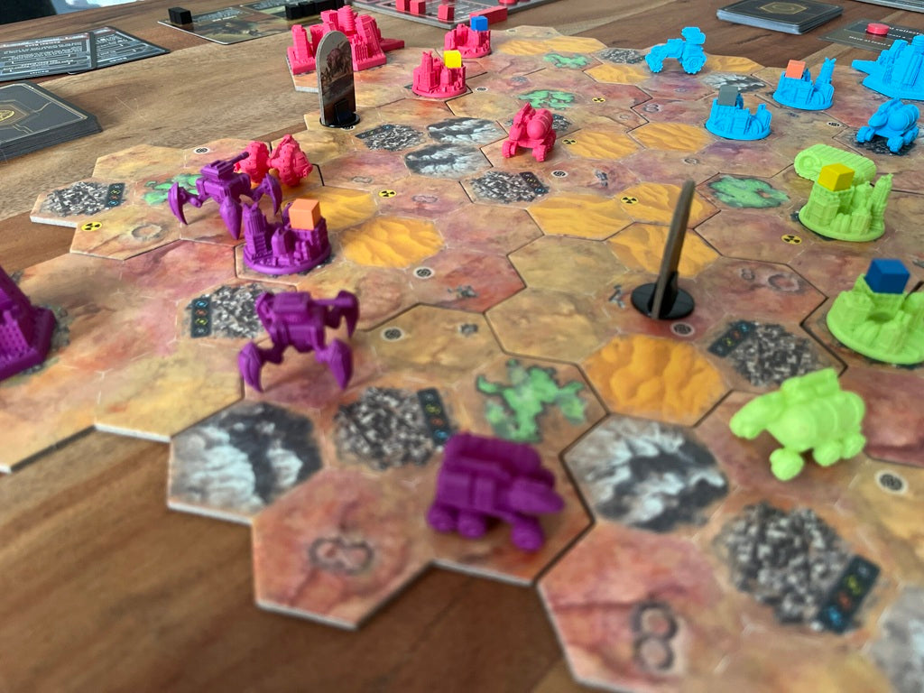 A close-up of Rise of the Wastelands Deluxe Kickstarter Edition by Wulfhorn Games shows hex tiles, colorful plastic figures, and yellow, blue, orange, and purple cubes on a wooden table. Some tiles display numbers and black tokens.