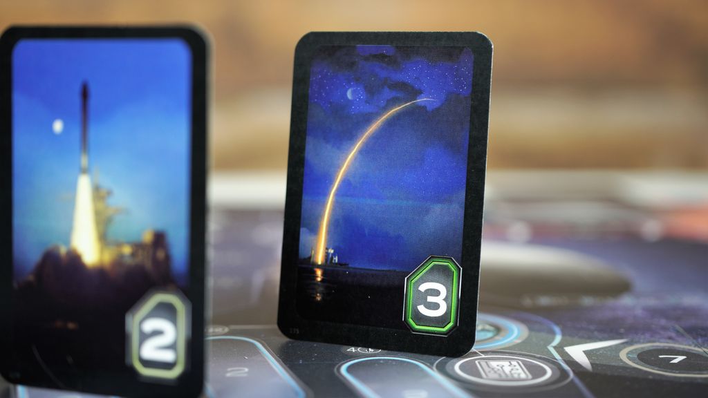 A close-up of two game cards from Rocketmen by Phalanx: one card shows a rocket launching at night with a glowing arc and green hexagon marked 3, while the blurred card depicts another launch with the number 2.