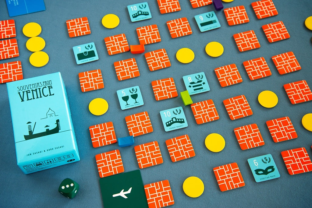 The Oink Games board game Souvenirs from Venice is set up on a gray surface with orange and blue square tiles for collection, yellow round tokens, a green die, and a light blue box.