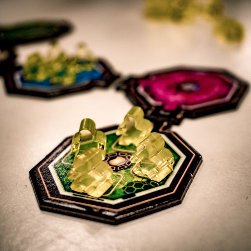 A close-up of translucent yellow game pieces on colorful hexagonal tiles from Ludonaute's Space Gate Odyssey, highlighting the space colonization theme with a sharp focus on the foreground tile and blurred background elements.