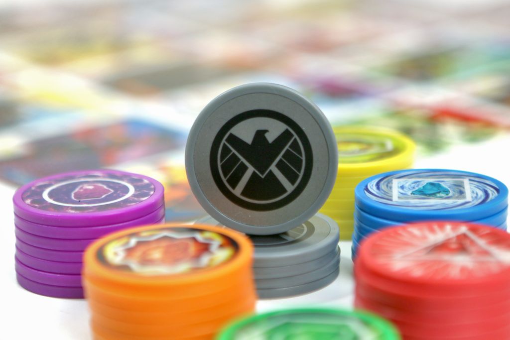 A close-up of vibrant game tokens stacked in piles, with a gray token featuring a black eagle emblem upright in the center, captures the strategic play and colorful components of Splendor: Marvel by Space Cowboys.